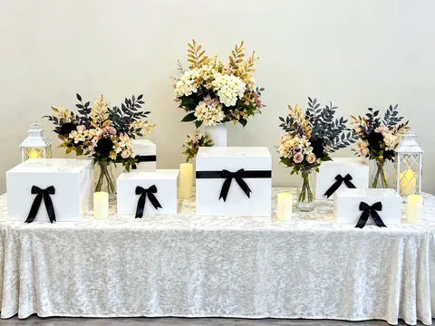 Dessert table styled with white display boxes tied with black ribbon bows, cream and gold floral arrangements, lanterns and candles on a velvet tablecloth
